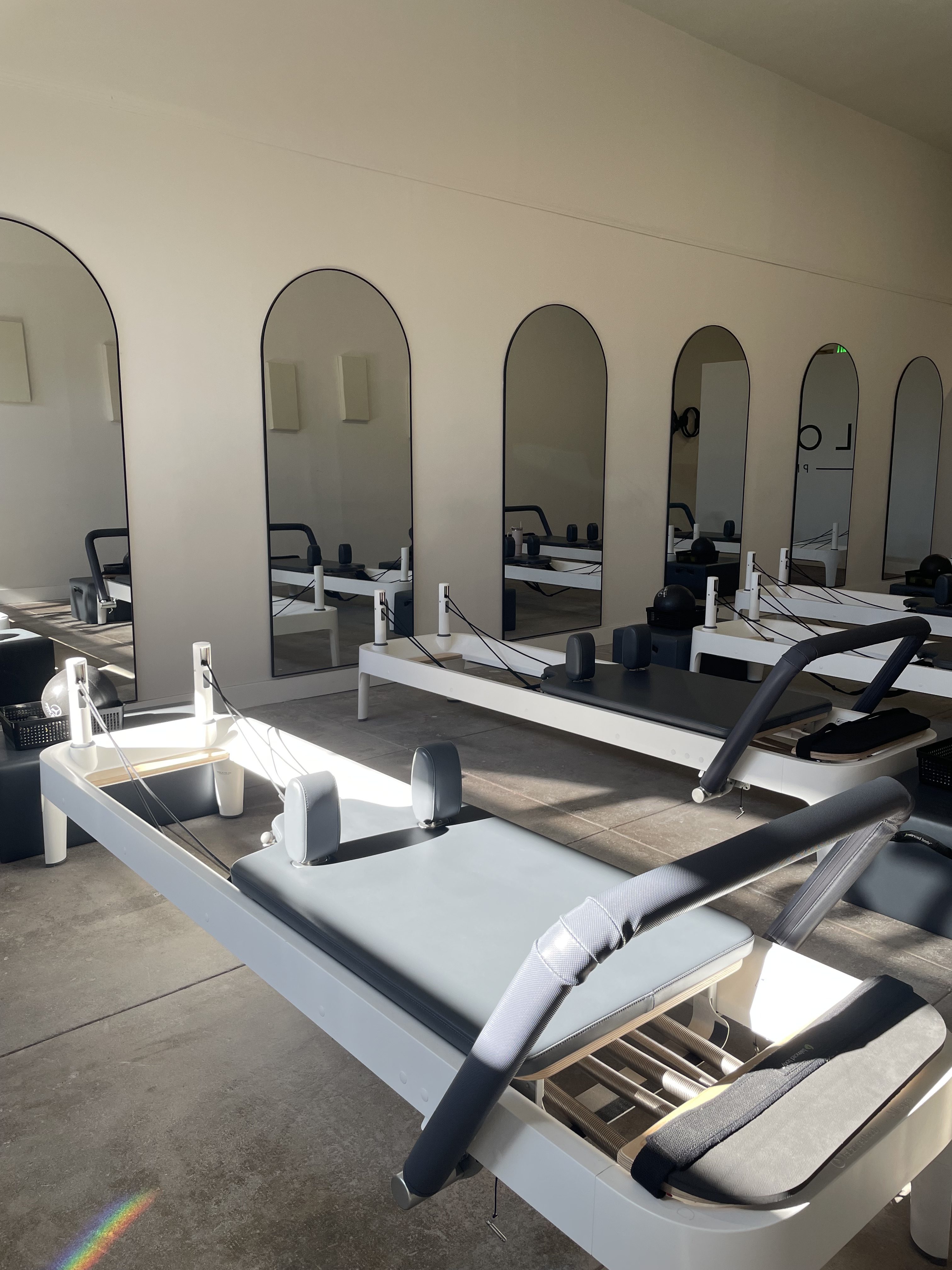 Interior view of a Pilates studio featuring multiple reformer machines arranged neatly, with large mirrors on the wall reflecting the setup.